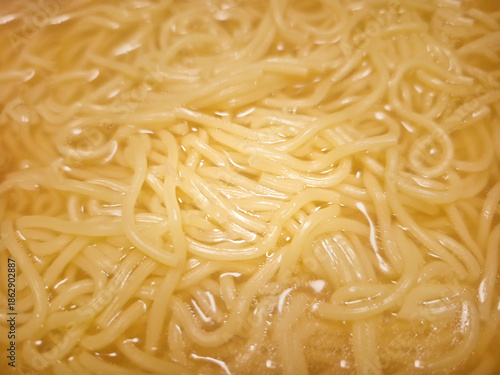 An extreme close-up of soft, yellow egg noodles submerged in a warm, clear savory broth, highlighting the slippery texture and light reflections of the soup.