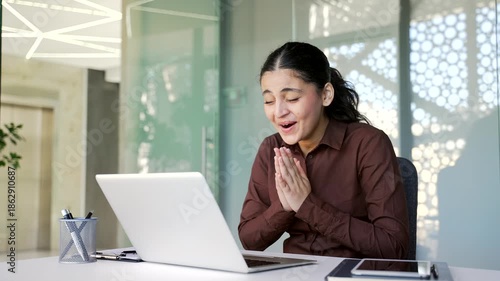 Happy excited young businesswoman reading great news on laptop computer while sitting at desk at workplace in business office. Joyful woman celebrates success, rejoices at victory, smiles. 