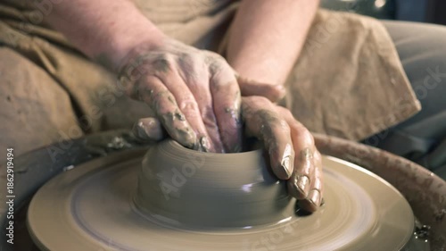 A craftsperson shapes clay on a pottery wheel, their hands covered in wet clay. The spinning wheel creates a smooth, symmetrical form, showcasing the skill and creativity involved in pottery making.