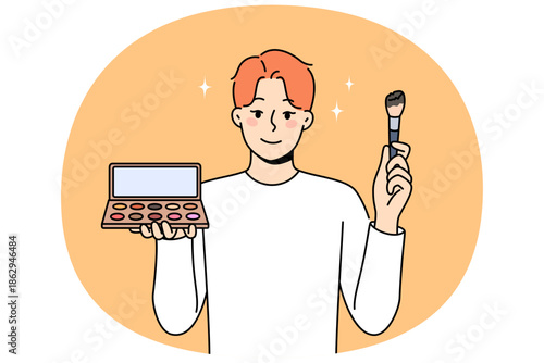 Non-binary man applies makeup and skin shadow, holding mirror and brush to apply powder. Non-binary guy uses cosmetics, wanting to become like girl, for concept of gender identity and tolerance