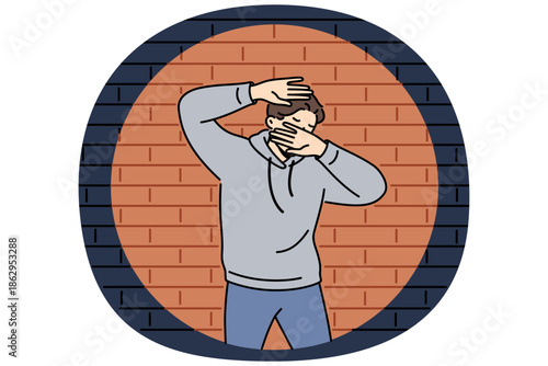 Man criminal in light of police lantern stands near brick wall and covers face with hands. Young guy criminal who committed robbery or murder was cornered after pursuit and investigation