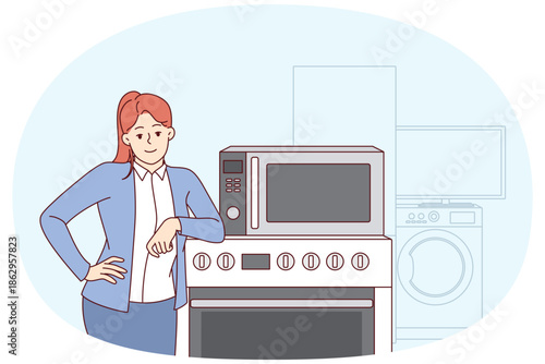 Woman selling household appliances stands near oven and washing machine and looks at camera. Girl smiles and rejoices in purchasing new home appliances with IOT wifi synchronization technology