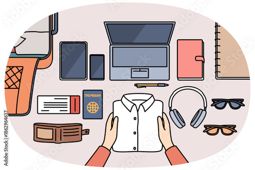 Personal items for business travel and suitcase for luggage are laid out on table near passport for customs control. Hands of man preparing clothes and gadgets for flight on business trip