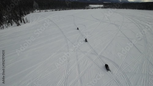 Tourists ride snowmobiles across the wide, frozen surface of Murray Lake in the Yukon wilderness, surrounded by boreal forest and snow-dappled mountains on a winter day