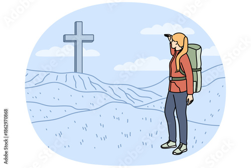 Woman pilgrim stands near large christian cross on hills during hike to sites of ancient cities. Pilgrim girl with big tourist backpack behind back looking at giant crucifix on horizon