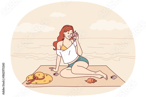 Woman sits on beach and listens to sound of sea from shell, enjoying summer vacation at tropical resort. Tourist girl sunbathes on beach of paradise island in ocean during weekend.
