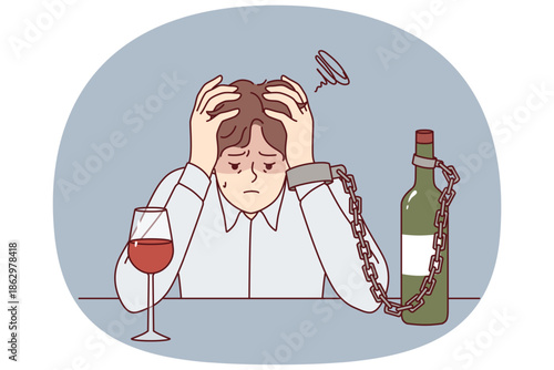Man suffering from alcohol addiction is chained to bottle of wine, sitting at table clutching head. Guy with liquor addiction fell into depression after being betrayed by girlfriend