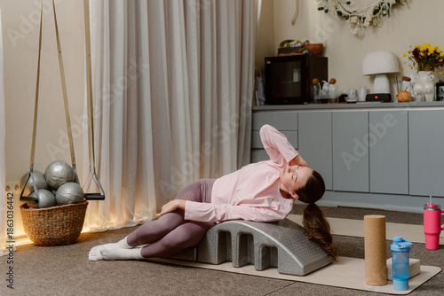 фотографія A young woman rests on a workout prop in her serene home gym, engaging in a gentle stretch while enjoying a moment of calm