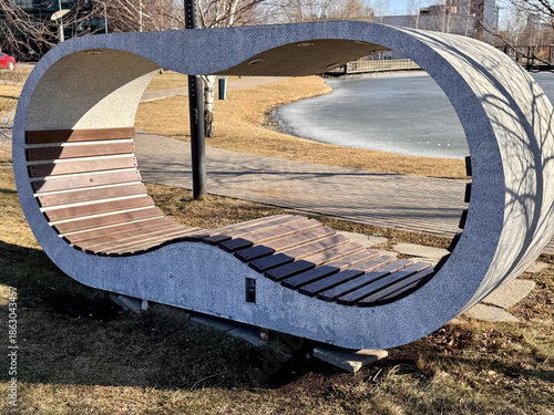 Elegant Lakeside Seat, Artistic Wooden Bench Near Water, Modern Curved Wooden Seat By Tranquil Lakeshore, Sculptural Wooden Bench With Flowing Form Located On Grassy Lakeside Bank