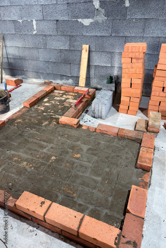 Fresh brickwork of a heating stove with a bench base. Laid out of brick on a concrete screed. Bricks and tools nearby.