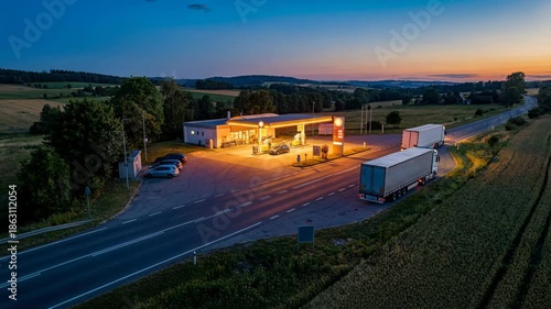 Illuminated Gas Station At Dusk With Trucks On Road Surrounded By Fields Under A Twilight Sky