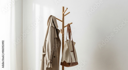 A minimalist wooden coat rack standing against a white wall, holding a beige trench coat and a canvas tote bag in a bright, sunlit room