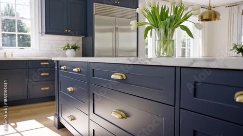 Bright kitchen featuring dark blue cabinets brass hardware white marble countertops a stainless steel refrigerator and white tulips