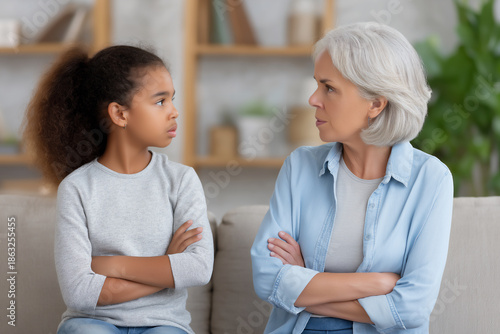 African American girl and senior woman sitting on a couch, facing each other with crossed arms, expressing disagreement in a cozy living room setting, showcasing generational conflict