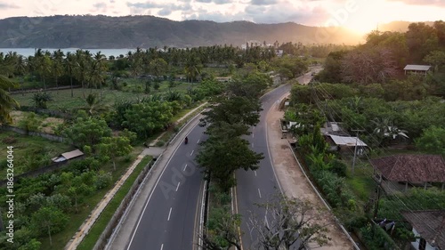 Aerial view of a winding coastal road with motorcycles driving through lush green scenery with palm trees and mountains during a beautiful golden sunset, showing the beauty of island life