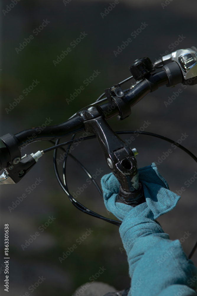 Fototapeta premium Gloved hand on bicycle handlebars gripping brake lever, blue glove and cable details visible, close framing shows rubber grip and lever texture, rocky trail and dim dusk light
