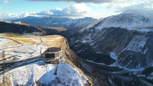 Aerial flight near Friendship Arch on Georgian Military Road. 4K horizontal drone footage shows the iconic monument surrounded by massive Caucasus mountains with snow-covered peaks in clear weather.