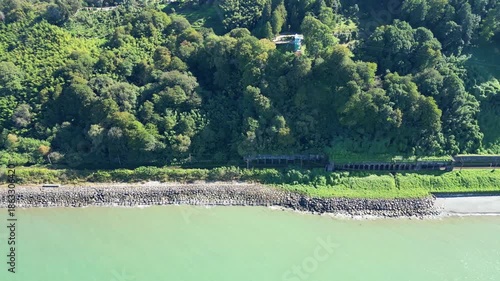 Aerial flight from sea along evergreen botanical garden in Batumi. 4K horizontal drone footage shows lush terraced greenery near railway tracks and coastline in clear sunny weather.