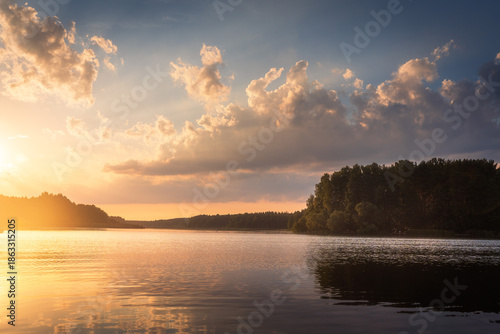Amazing sunrise over lake with colorful clouds at the horizon. Forest on the lake side. Sunny morning in wild nature.
