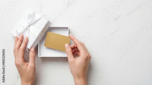Fotografía A top-down flat lay features well-manicured hands placing a blank, metallic gold gift card into a minimalist white gift box tied with a silk ribbon