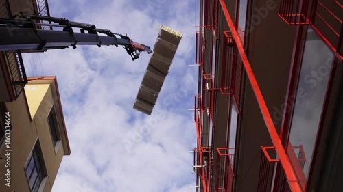 A construction crane lifts insulated roofing panels. A hydraulic truck crane precisely moves large, shrink-wrapped stacks of insulated sandwich panels between closely spaced residential buildings.