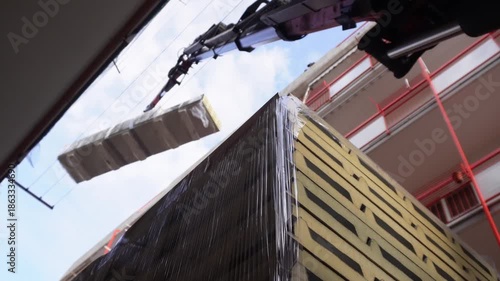 A construction crane lifts insulated roofing panels. A hydraulic truck crane precisely moves large, shrink-wrapped stacks of insulated sandwich panels between closely spaced residential buildings.