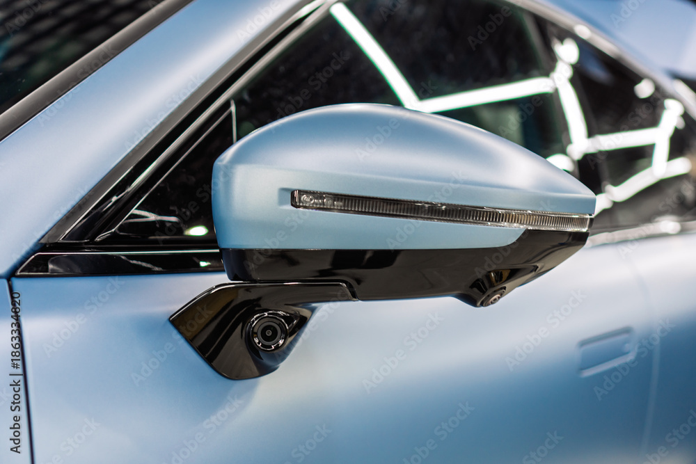 Fototapeta premium Car side mirror with integrated camera blue car, cam for driver safety and visibility. Car side view camera system integrated into rearview mirror, blind spot detection, 360 visibility. Camera 360. 