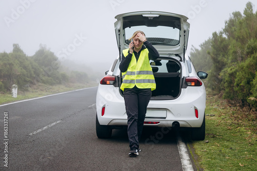 Woman in reflective vest stands beside broken car on roadside, expressing frustration with vehicle issues in foggy weather