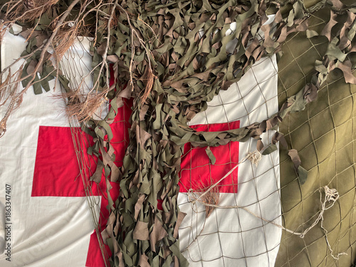 Camouflaged medical tent wall with a red cross symbol, covered by netting and natural elements. Concept of military medicine, battlefield aid, emergency care, humanitarian support, and protection.