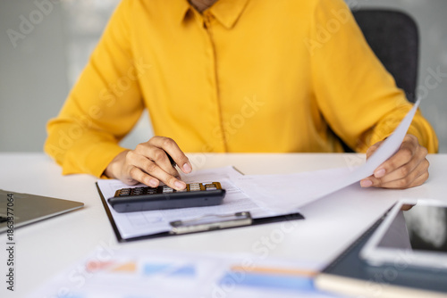 Person wearing a yellow shirt using a calculator and reviewing paperwork for financial planning, accounting, and preparing taxes in a professional office setting