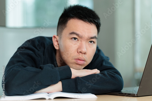 Obraz na plátně A weary Asian student rests his head on his crossed arms at a desk with an open textbook and laptop, ideal for student fatigue, academic stress, or study exhaustion concepts