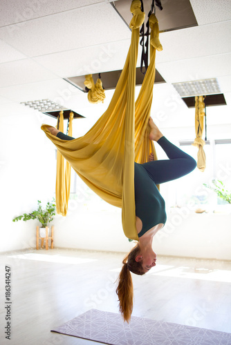 A woman hangs upside down from a yellow aerial yoga hammock in a studio. Sunlight fills the space, and yoga mats are seen on the floor. The atmosphere feels focused and active