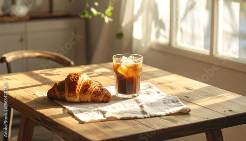 Cozy home cafe dining table with a linen cloth, iced coffee, and a fresh croissant, morning shadows with analog film grain.