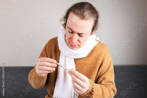 Fényképezés Sick man with red nose and scarf checking body temperature with thermometer