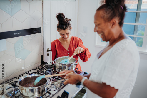 Photos Happy young woman and her mother cooking together in a modern kitchen, stirring pots on a gas stove and sharing a joyful moment while preparing a meal at home