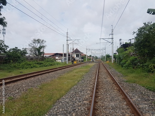 Railway tracks leading toward a small station building in the distance. Infrastructure and overhead power lines on a bright cloudy day. Train tracks and green nature leading to a destination.