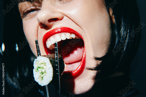 Young woman with short black hair enjoying sushi, vibrant red lips, and expressive emotions against a dark background, capturing a lively culinary moment.