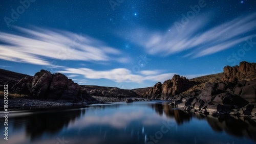 Long exposure photography of a rocky shoreline under a starry sky with wispy clouds reflecting in the calm water