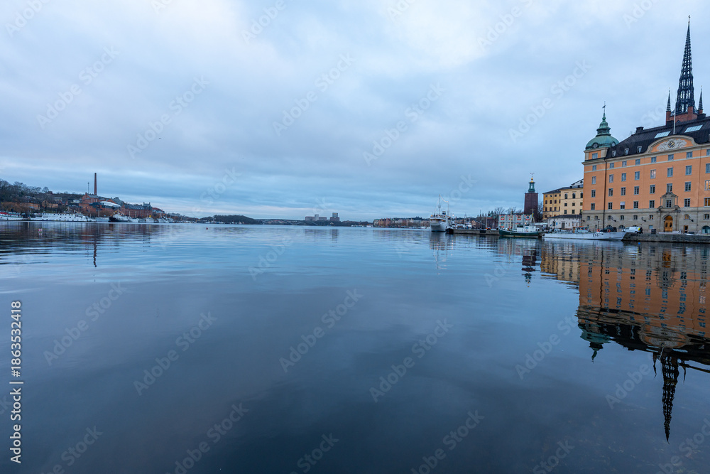 Obraz premium Calm winter reflections across the waterfront in Stockholm, Sweden, with the historic skyline of Gamla Stan and the soaring spire of Riddarholmen Church mirrored on the glassy harbor.