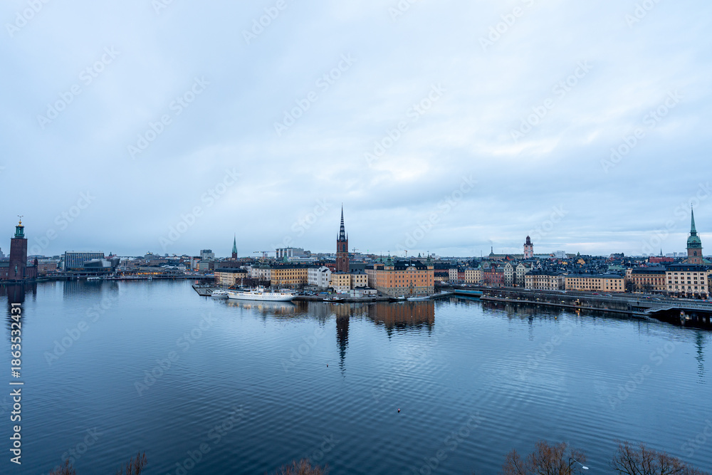 Fototapeta premium Stockholm skyline across Riddarfjärden with Riddarholmen Church and Stockholm City Hall reflected on calm water under an overcast sky, Sweden.