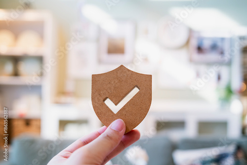 Hand holding wooden shield icon with check mark in modern home interior, representing home security, insurance protection, trust assurance, safety verification, quality controlใ