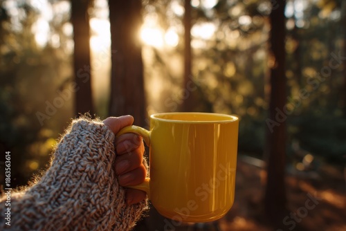 Hand holding a yellow mug outdoors in a forest at sunrise
