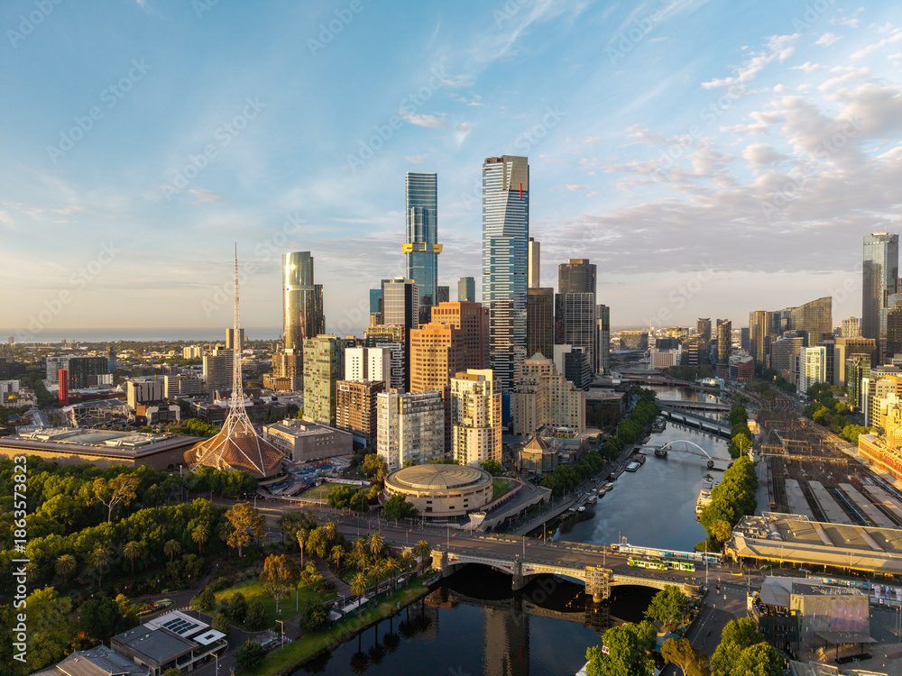 Naklejka premium Melbourne's expansive urban landscape featuring towering skyscrapers, the winding Yarra River, bridges, and green spaces bathed in golden light.