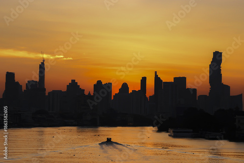 Boat glides along river against backdrop of city skyline at sunset, casting warm orange glow over water and silhouetting tall buildings