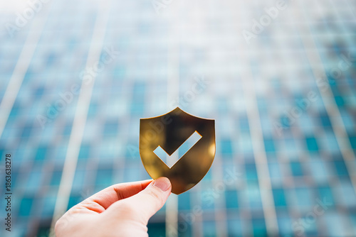 Hand holding a golden shield icon with check mark symbol on blur modern glass building background, representing cybersecurity, trust, data protection, online privacy, and secure technology concept.