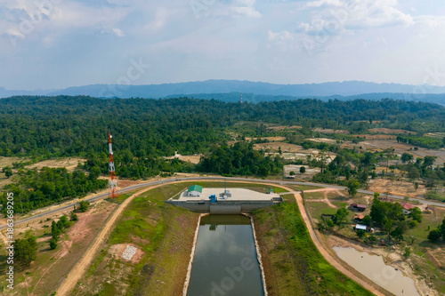Aerial perspective shows a modern water control facility with a green-roofed utility building and a tall communications tower beside a canal, surrounded by grassy embankments, scattered houses, and