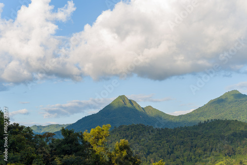 Lush green forested mountains rise beneath a sky filled with large, billowing clouds near Dien Bien Phu, Vietnam. Soft sunlight highlights the landscape, creating a fresh and expansive natural scene.