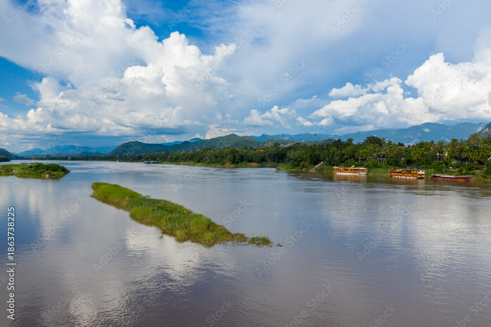 Obraz premium Large cumulus clouds tower above the Mekong River as it winds past grassy islets and tropical banks near Luang Prabang, Laos. Wooden boats line the lush shoreline, with distant mountains providing a