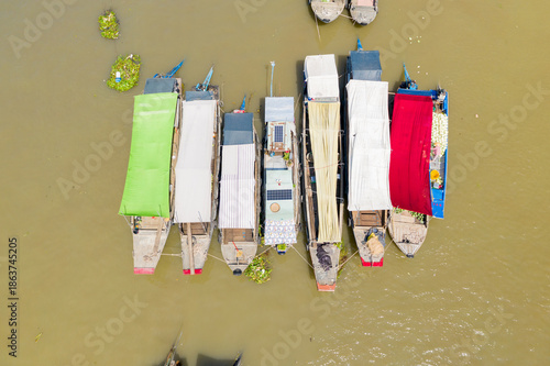 Top-down view features a row of covered market boats moored side by side on the muddy river at Cai Rang floating market in Can Tho, Mekong Delta, Vietnam. Distinctive tarps in green, white, and red
