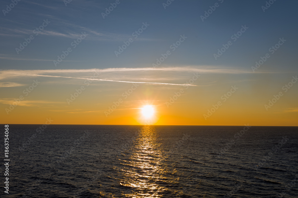 Fototapeta premium The sun sets low above the calm Ionian Sea, casting a luminous golden reflection across gentle waves. Subtle cloud streaks and a gradient sky create a tranquil, minimal seascape.